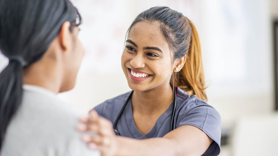 A smiling nurse in scrubs places a comforting hand on a patient's shoulder.